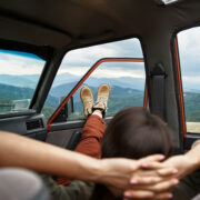 Person laying back with their hands behind their back with their feet out the window of a parked car in a mountain valley