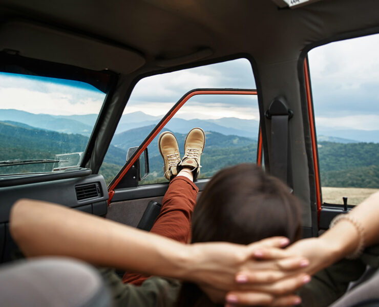 Person laying back with their hands behind their back with their feet out the window of a parked car in a mountain valley