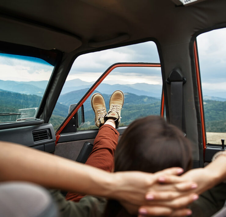 Person laying back with their hands behind their back with their feet out the window of a parked car in a mountain valley
