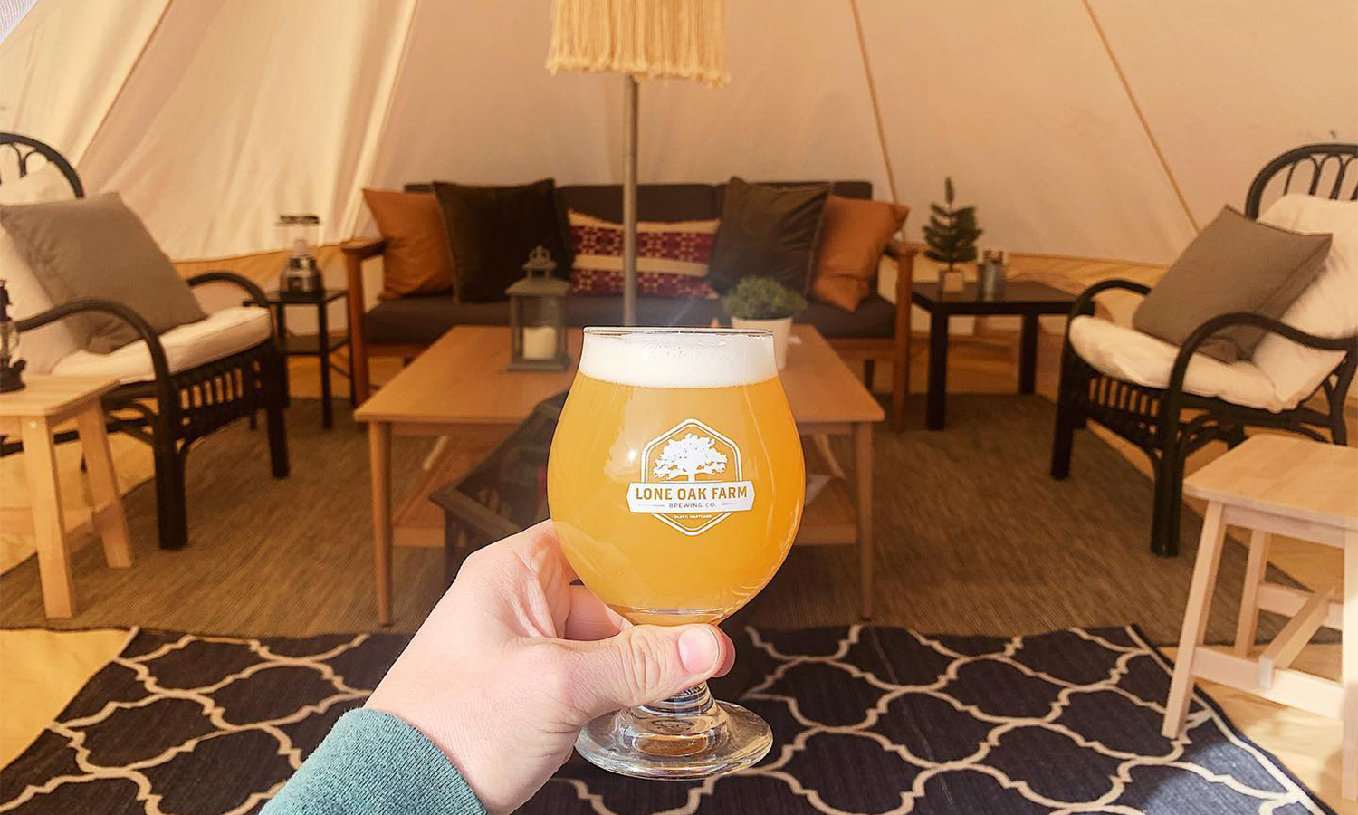 Woman hand holding up glass filled with beer in front of tent with chairs and rugs